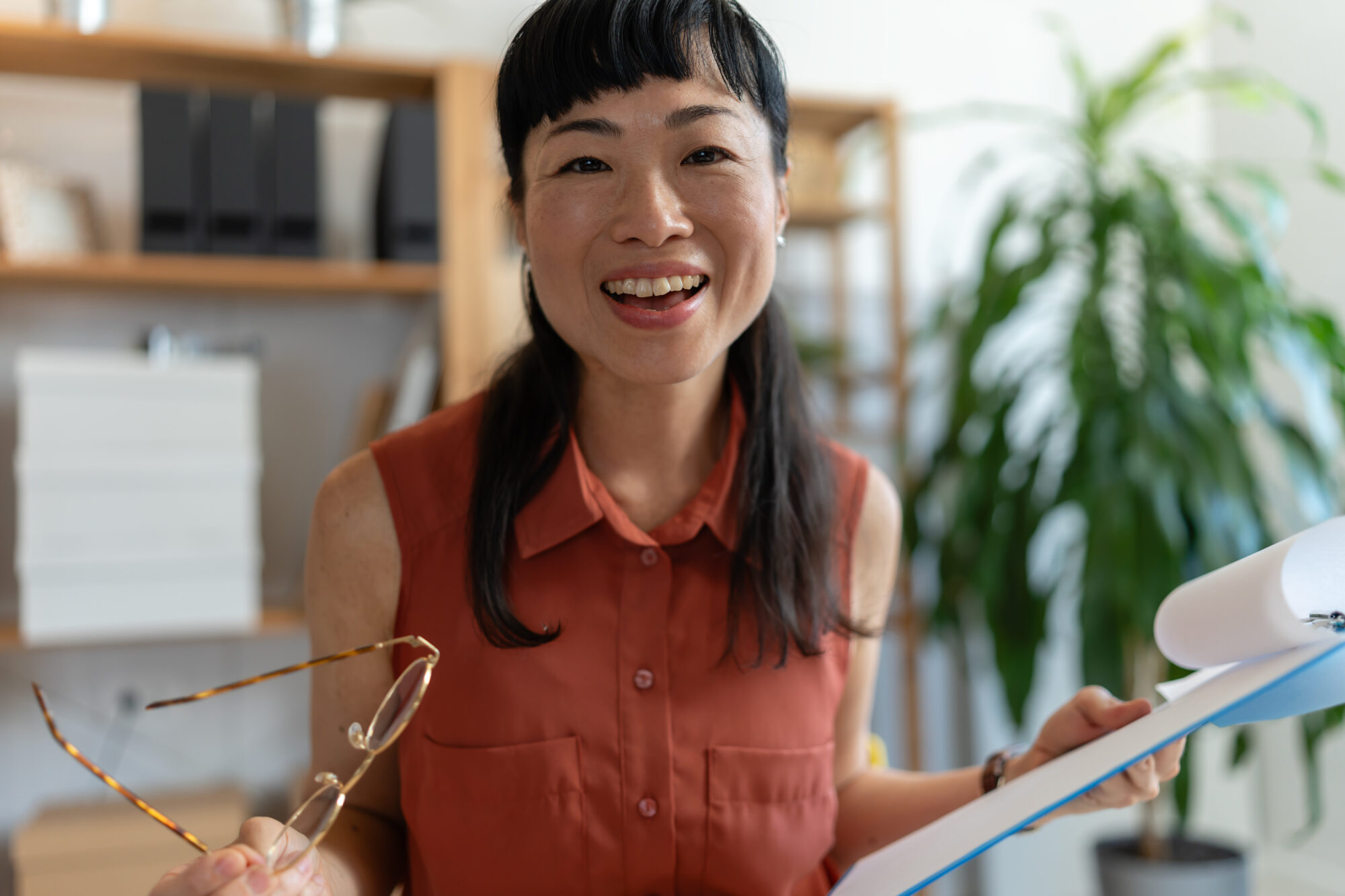 Asian woman small business owner in her home office, smiling and gesturing while holding documents, appearing engaged in a friendly discussion or presentation